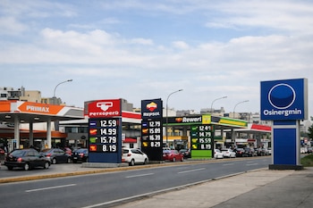 Fila de estaciones de servicio en una avenida de Lima con carteles de precios de combustibles de Primax, Petroperú, Repsol y Rewel, y un cartel de Osinergmin.