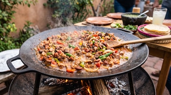 Un disco metálico sobre brasas de madera contiene pedazos de carne y vegetales en cocción, junto a una mesa con tortillas y salsas.