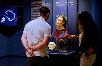 Hombre y mujer observan un busto de reconstrucción facial con corona de hojas, un cráneo humano y una pantalla con tomografía computarizada de cráneo
