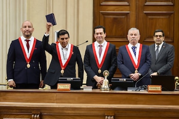 Cinco hombres con traje, corbata y bandas ceremoniales. Uno de ellos levanta un libro oscuro. Hay un escritorio de madera, micrófonos y una campana