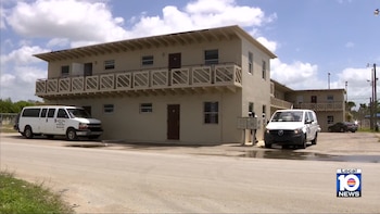 Dos camionetas blancas estacionadas frente a un complejo de apartamentos de color beige de dos pisos con balcones. El cielo está parcialmente nublado