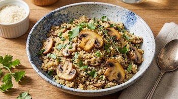 Plato hondo de cerámica con quinoa, champiñones, perejil y tomillo. Se ven un recipiente blanco con queso rallado y una cuchara sobre un paño de lino.
