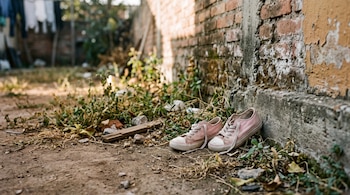 Un par de zapatillas infantiles de tela rosa sobre tierra junto a un muro de ladrillo y maleza.