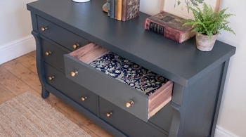 Cómoda oscura con un cajón abierto de interior floreado, libros antiguos y una pequeña maceta con helecho sobre la tabla superior.