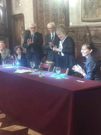 Angela Pradelli sonríe mientras recibe un galardón. Dos hombres de traje de pie y tres personas sentadas aplauden. Fondo con paneles de madera tallada y tapices