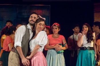 Grupo de actores en un escenario con vestuario de época; un hombre y una mujer en primer plano sonríen, otra mujer sostiene una regadera verde