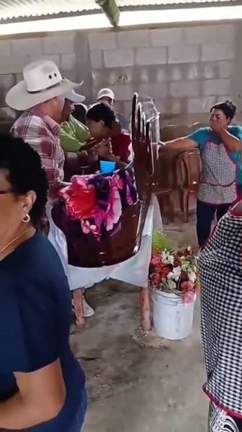 Personas reunidas en una habitación de concreto alrededor de un ataúd de madera con flores al lado