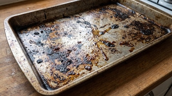 Bandeja rectangular de metal con residuos oscuros de grasa quemada sobre una superficie de madera en una cocina.