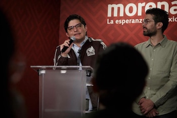 Un hombre con gafas y chaqueta habla en un micrófono en un atril transparente. Otro hombre con barba y gafas está de pie a su lado. Fondo rojo con texto Morena