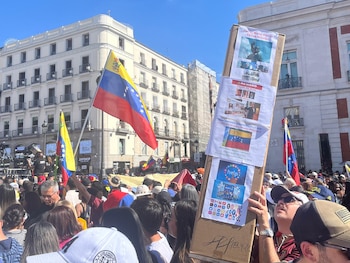 Multitud en una plaza con banderas venezolanas ondeando, edificios históricos de fondo y pancartas con mensajes políticos sostenidas por manifestantes
