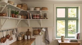 Estantes blancos con cestas, cuencos, libros, plantas. Encimera de madera. Frascos de comida, tablas de cortar, utensilios, ollas de cobre. Ventana de marcos verdes al jardín. Jarra y libros.