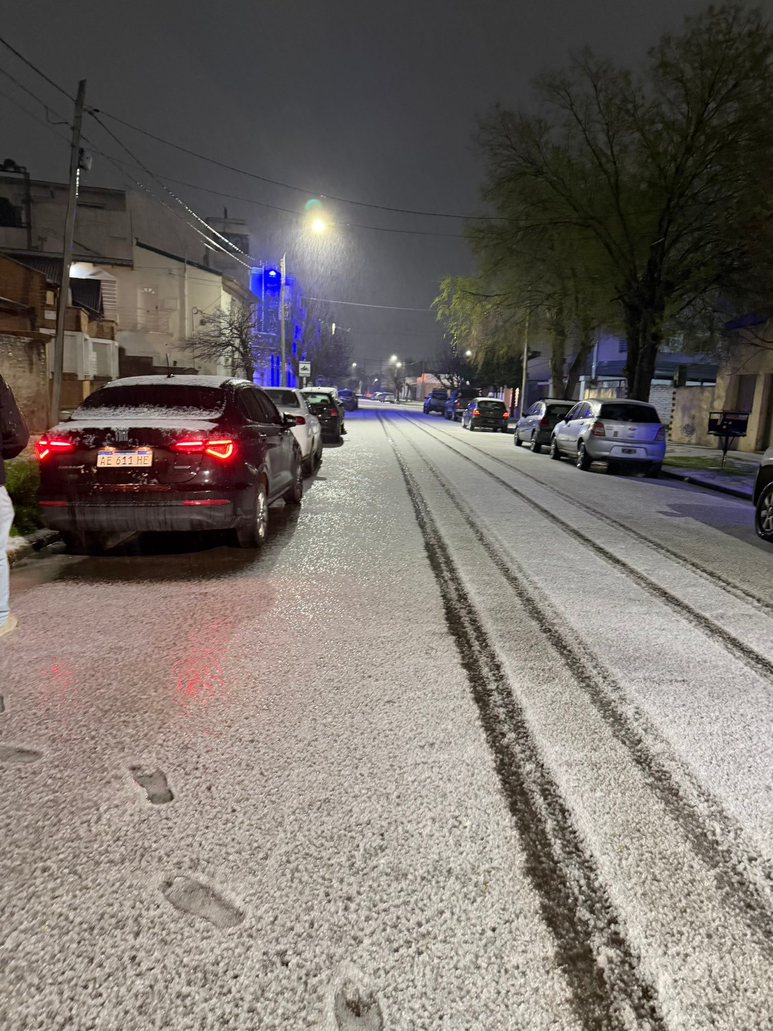 Varios automóviles permanecen estacionados en una calle residencial cubierta por una capa de nieve con marcas de neumáticos durante la noche.