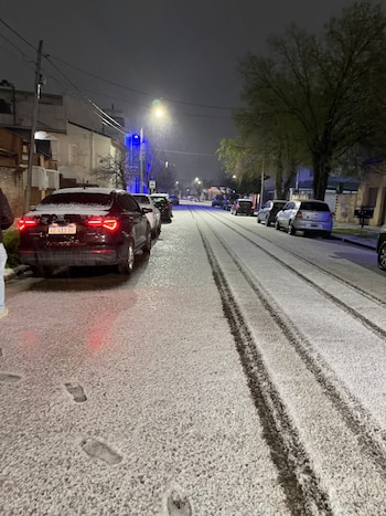 Calle nocturna cubierta de nieve con marcas de ruedas, pisadas en el suelo y automóviles estacionados junto a las aceras
