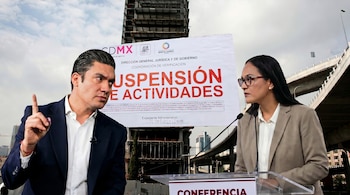 Hombre y mujer en conferencia frente a un letrero oficial que anuncia 'Suspensión de Actividades'. Un edificio en construcción y un puente elevado de fondo.