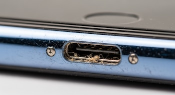 Primer plano macro del puerto de carga USB-C de un teléfono móvil azul con mucha pelusa y suciedad dentro del conector. Se ven dos tornillos pequeños.