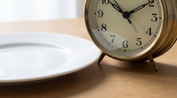 Un reloj despertador clásico dorado y un plato blanco vacío reposan sobre una mesa de madera clara, con iluminación suave.