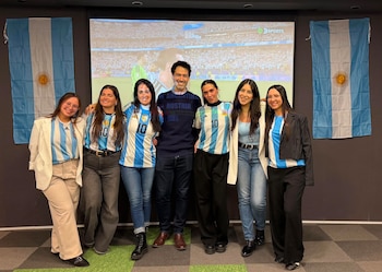 Siete personas, seis mujeres y un hombre, posan frente a una pantalla con un partido de fútbol. Detrás, dos banderas argentinas y suelo de cuadros