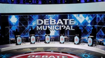 Seis siluetas humanas tras podios iluminados en un escenario con una pantalla azul de fondo con el texto Debate Municipal.