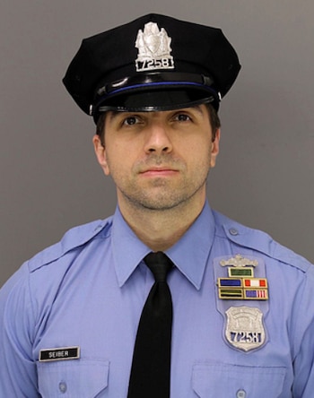Un hombre uniformado de policía viste camisa azul, corbata negra, gorra oficial y placa dorada con el número 72581