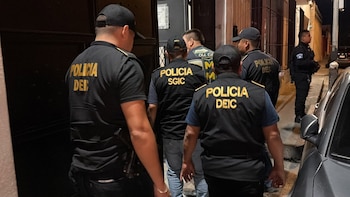 Varios policías con uniformes negros y letras doradas, algunos de espalda, otros de perfil. Se observa un edificio oscuro y un coche gris estacionado en la calle