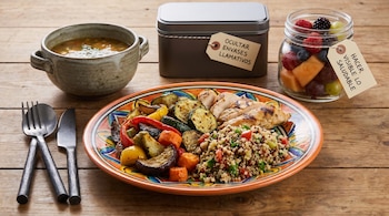 Composición en mesa de madera con plato de pollo, quinoa y verduras, cuenco de sopa, cubiertos de metal, caja opaca y tarro de cristal con frutas variadas.