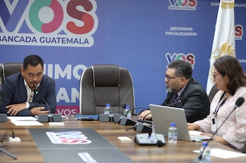 Tres personas sentadas en una mesa con micrófonos, botellas de agua y un portátil. Fondo azul con logos de VOS Bancada Guatemala y una bandera blanca