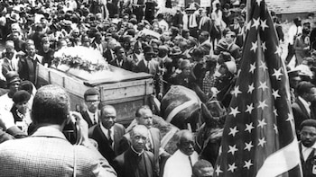 Imagen en blanco y negro de la procesión fúnebre de Martin Luther King Jr. Se ve un ataúd en una carreta tirada por mulos y una bandera de EE.UU. entre una gran multitud