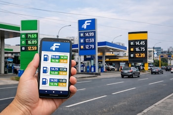 Una mano sostiene un teléfono móvil con una aplicación de comparación de precios de combustible. Al fondo, tres estaciones de servicio con sus carteles de precios.