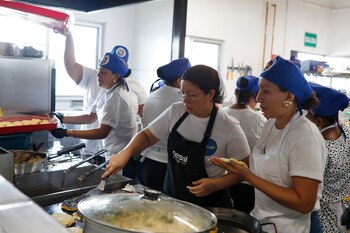 Una mujer sonriente con gorro y delantal blanco con un logo de comida, sostiene una bandeja negra con tres platos de comida. Al fondo, personas y pancartas
