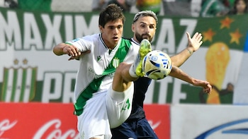 Dos futbolistas en acción; uno con camiseta blanca y verde intenta controlar un balón con el pie, mientras otro de camiseta oscura lo marca