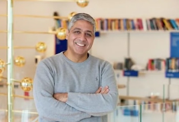 Un hombre con el cabello canoso sonríe con los brazos cruzados frente a estanterías con libros y una estructura dorada