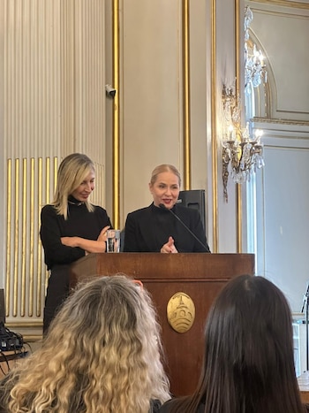 Una mujer de cabello rubio habla desde un podio de madera con un emblema, con otra mujer de pie a su izquierda y la parte trasera de la audiencia en primer plano