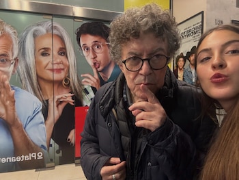 Un hombre con gafas y cabello rizado se lleva el índice a los labios junto a una mujer joven en un vestíbulo con carteles