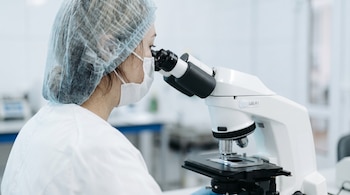 Mujer con gorro sanitario, mascarilla facial y bata blanca mirando a través de un microscopio de laboratorio con platina para muestra.