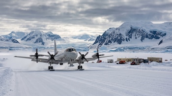 Avión de hélice blanco en una pista de nieve, con montañas nevadas, nubes y edificios modulares de campamento al fondo.