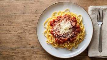 Un plato con fideos largos amarillos, salsa roja de tomate y queso blanco rallado. Una servilleta de tela y un tenedor plateado a la derecha sobre mesa de madera