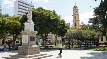 Estatua de piedra sobre un pedestal en una plaza pública con árboles, personas sentadas y la torre de una catedral al fondo.