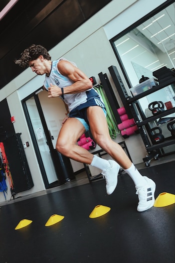 Hombre joven en ropa deportiva corre entre conos amarillos en un gimnasio. Al fondo hay bandas elásticas, rodillos de espuma y kettlebells