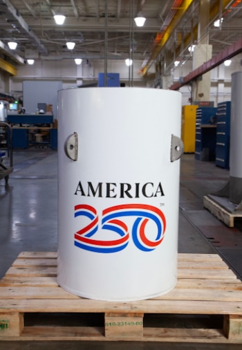 Cápsula del tiempo cilíndrica de color blanco con el logo "AMERICA 250" en rojo y azul, sobre un palé de madera en un entorno industrial