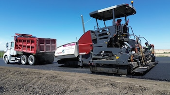 Un tren de pavimentación gris y rojo extiende asfalto en una carretera junto a un camión de volteo blanco con caja roja, bajo un cielo azul despejado