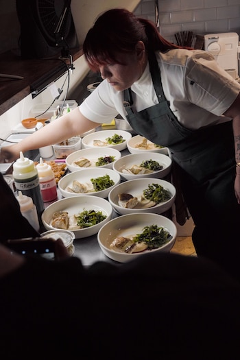 Una chef con pelo rojo y delantal oscuro emplata una fila de cuencos blancos con porciones de pescado, guarnición verde y puré blanco en una cocina industrial con botellas de salsa