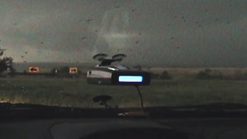 Vista desde el interior de un coche a través del parabrisas con gotas y partículas. Un dispositivo electrónico con pantalla azul está montado. El fondo muestra un campo verde bajo un cielo gris oscuro