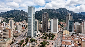 Edificios altos y modernos en una zona urbana junto a una avenida con tráfico, plazas con árboles y montañas al fondo.