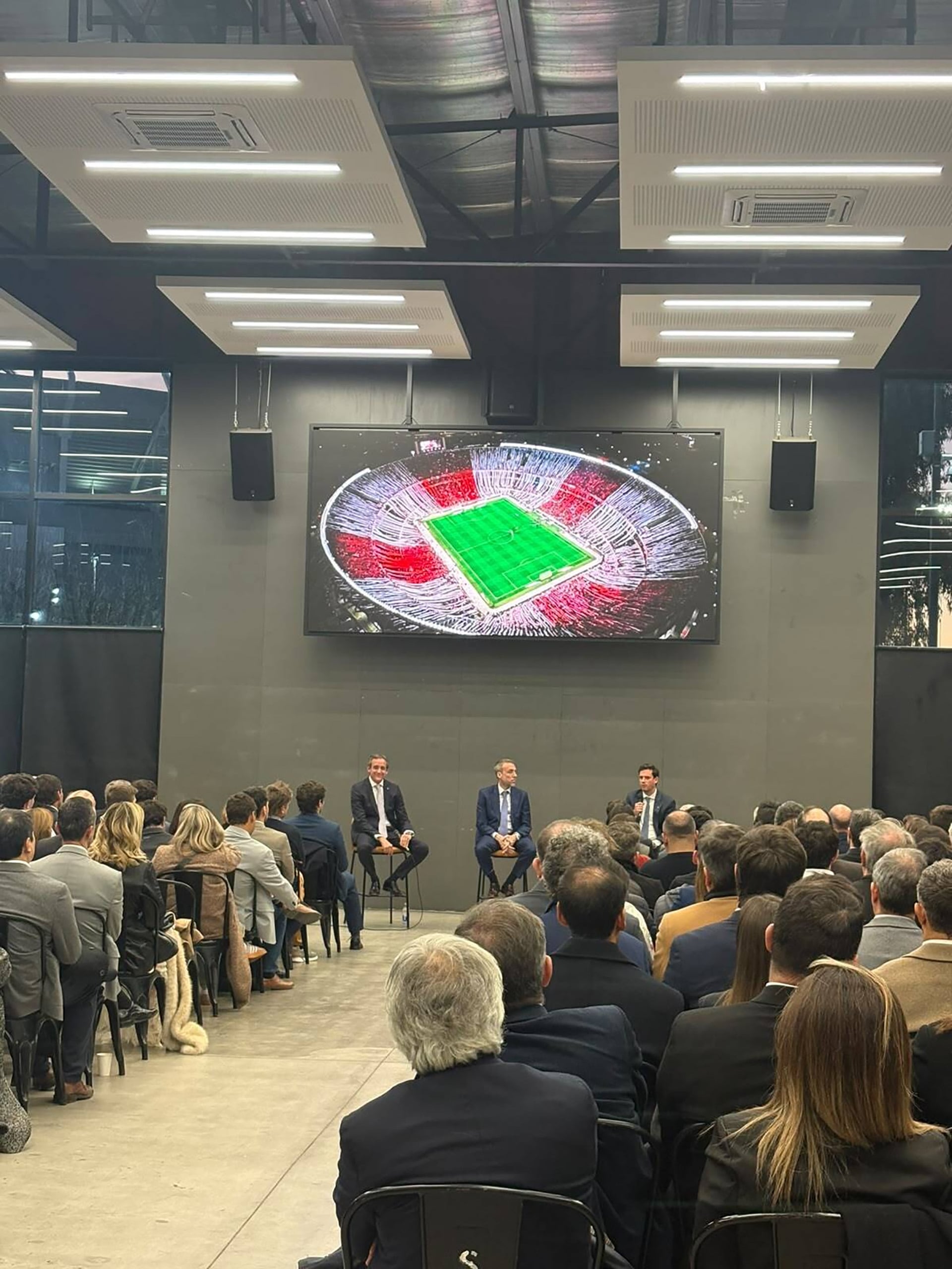 Tres panelistas dialogan durante un seminario en un auditorio mientras una pantalla gigante proyecta la vista aérea de un estadio de fútbol.