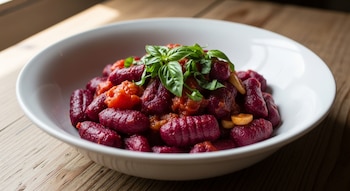 Plato hondo blanco con ñoquis de remolacha de color morado, salsa de tomate roja y hojas de albahaca verde, sobre una mesa rústica de madera.