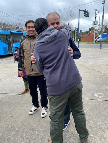 Un hombre con canas y un hombre con sudadera se abrazan en la calle. Otro hombre sonriente está detrás. Al fondo se observa un autobús azul y semáforos