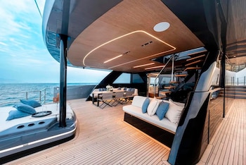 Cubierta de madera en una embarcación con sofás blancos, cojines azules, mesa con sillas, escaleras y vista al mar