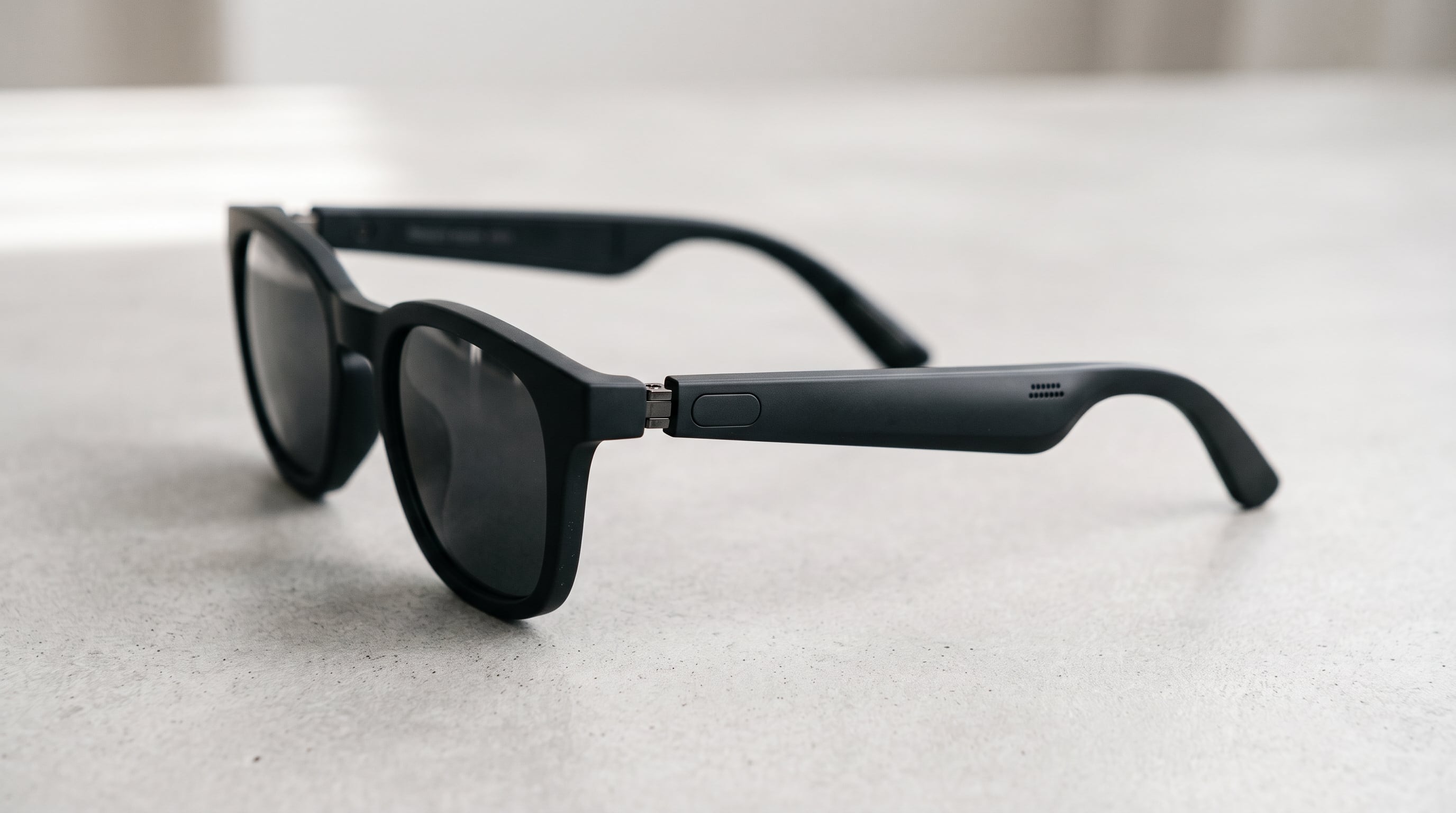 Unas gafas inteligentes de sol de color negro reposan sobre una superficie clara con un botón de control en la patilla. (Imagen Ilustrativa Infobae)