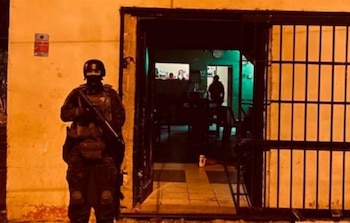 Agente de seguridad con uniforme táctico y rifle de pie junto a una puerta abierta de una casa de noche. Se observa la silueta de una persona en el interior