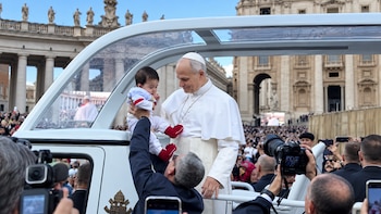 El papa vestido de blanco en un vehículo descubierto sostiene a un bebé que le acerca un guardia, rodeado de fotógrafos y público